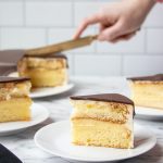 A photo of three plated slices of Boston Cream Pie on a marble surface, with a hand coming into frame to cut another slice from the rest of the cake in the background.