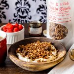 Cherry Cacao Granola: A lovely local collaboration! A photo of a wooden bowl of yogurt topped with Cherry Cacao Granola, with a bag of granola, a small ramekin of raspberries, and a cup of coffee resting beside the bowl on a wooden surface.