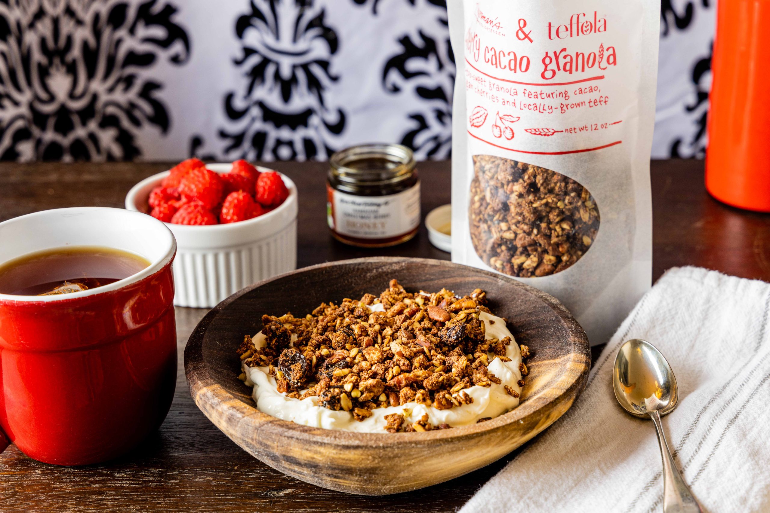 A photo of a wooden bowl of yogurt topped with Cherry Cacao Granola, with a bag of granola, a small ramekin of raspberries, and a cup of coffee resting beside the bowl on a wooden surface.