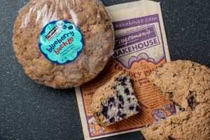 Two Blueberry Buckles with a Bakehouse pastry bag underneath. One of the Buckles is still wrapped, while the other is unwrapped and has a slice cut from it.