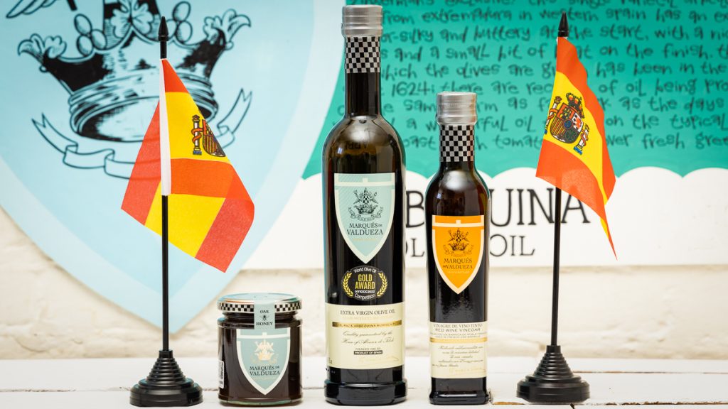 a photo of a bottle of Marques de Valdueza olive oil, vinegar and honey in front of a painted poster flanked by 2 spanish flags
