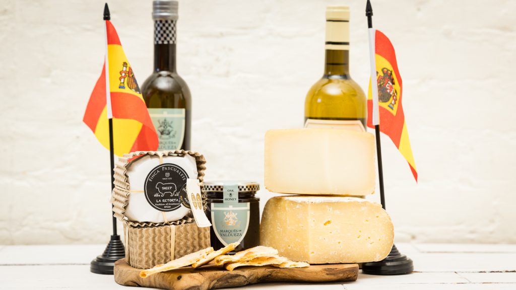a photo of 3 spanish cheeses, a jar of honey, some tortas, a bottle of wine and olive oil and 2 spanish flags