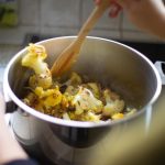 A pot of roasted cauliflower on a stove, with a hand coming into frame with a wooden spoon, stirring the cauliflower.