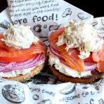 A sliced and toasted sesame bagel on a sheet of Zingerman's deli paper topped with whitefish salad, sliced red onion, lox, and more whitefish salad.