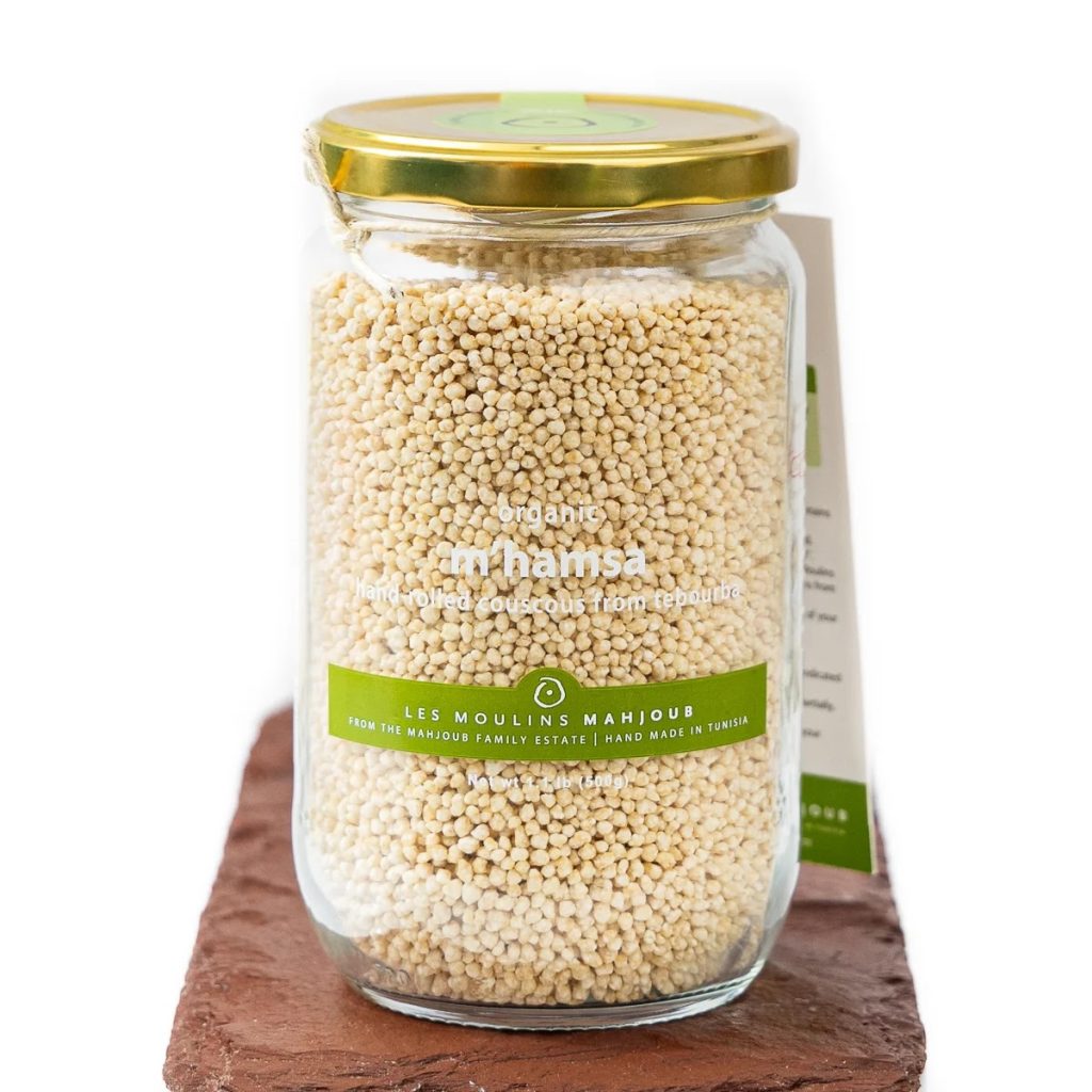A photo of a jar of Les Moulins Mahjoub's M'Hamsa Couscous. The jar is made of clear glass with a gold lid and is sitting on a brown slate serving board.
