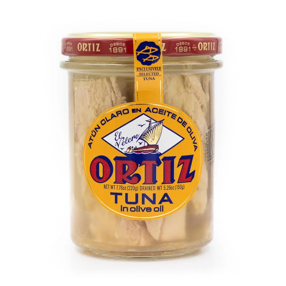 A photo of a jar of Ortiz Tuna in Olive Oil. The jar is made of clear class with a red lid and a deep yellow label.