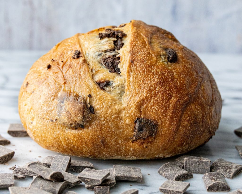 A photo of a loaf of Chocolate Chunk Sourdough on a marble surface with pieces of chocolate surrounding it. 