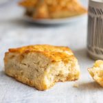 Ginger Scones Get Better Still with Cultured Butter A photo of a Ginger Scone on a light gray wooden surface with a plate of more scones and a coffee mug blurred in the background.