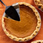 Exceptional Pumpkin Pie from the Bakehouse A top-down photo of a whole pumpkin pie with a slice taken out on an orange tablecloth, with a pie slicer resting in the empty space of the pie tin.