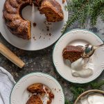 A top-down photo of a Gingerbread Coffeecake on a circulate white plate, with two plated slices and a dish of whipped cream next to it, and a sprig of pine peeking in from the top right corner.