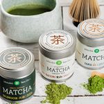 Introducing: Mizuba Tea Co. A photo of three tins of Mizuba Matcha on a white painted wooden surface, each with a small pile of powder in front of it, with a matcha whisk and ceramic matcha bowl with matcha inside it behind the tins.