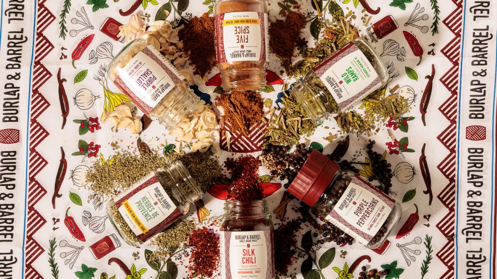 A top-down photo of six different Burlap & Barrel spice jars atop a Burlap & Barrel bandana, with the ground spices spread atop the fabric.