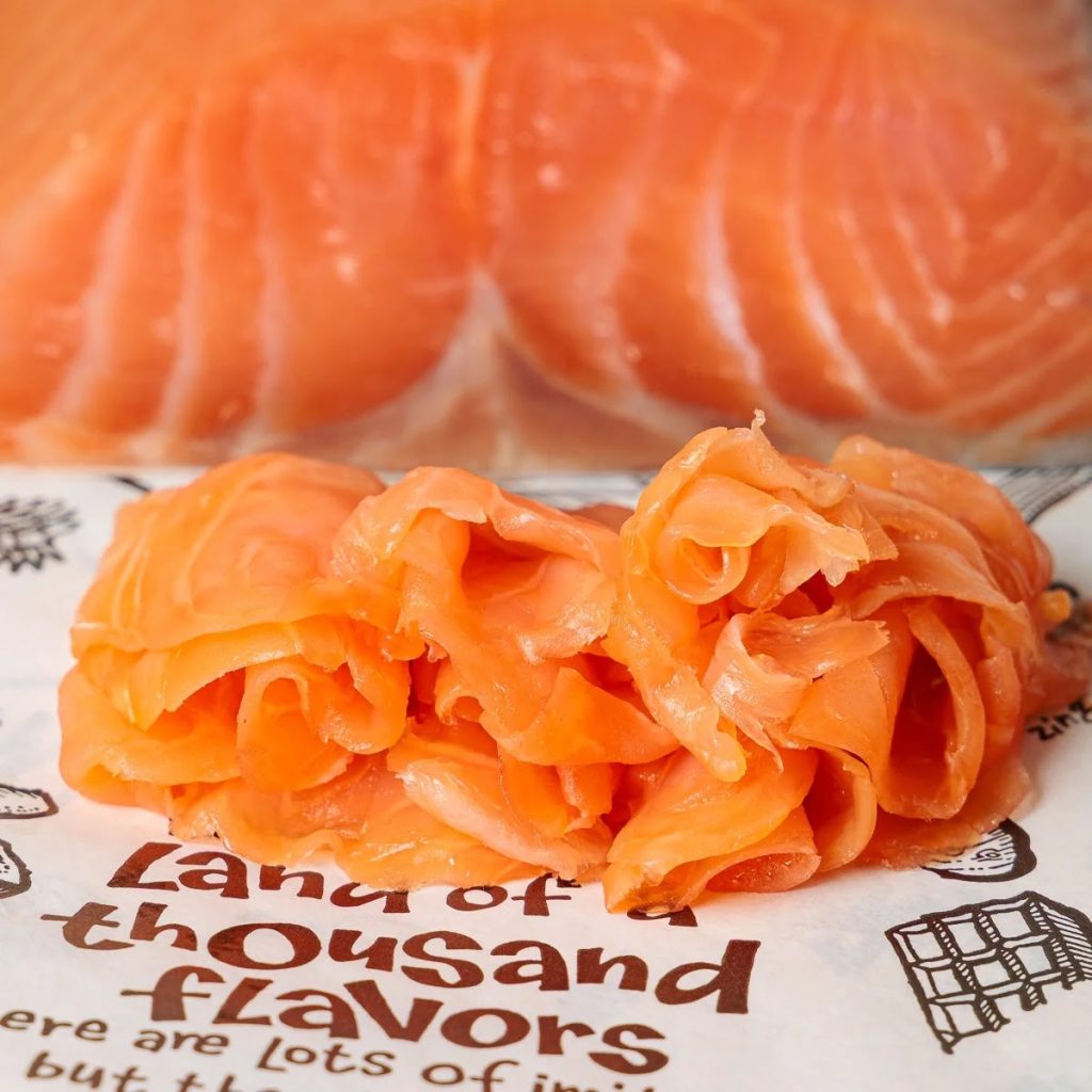 A photo of a pile of thinly-sliced smoked salmon resting on a sheet of Deli paper, with more unsliced smoked salmon in the background.