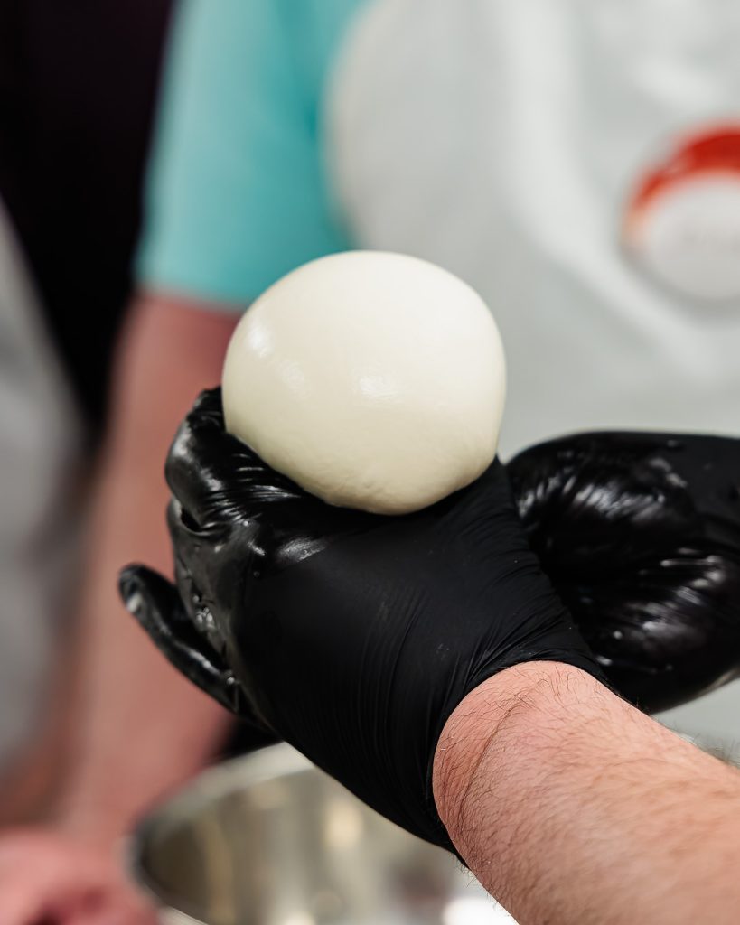 A photo of a set of gloved hands shaping a ball of mozzarella.