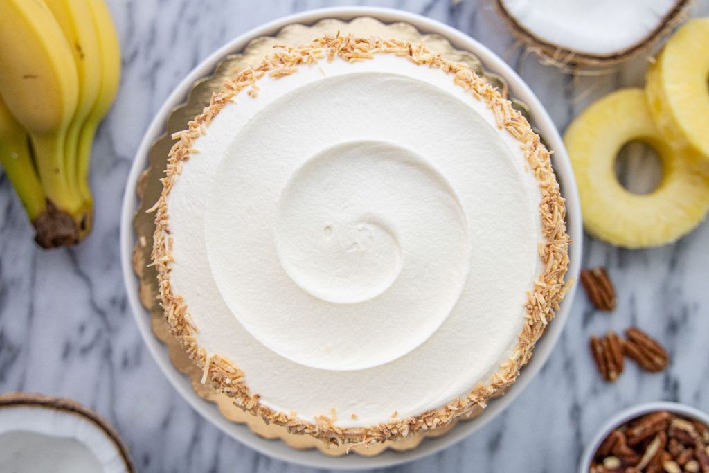 A top-down photo of a Hummingbird Cake on a marble surface, with a bunch of bananas, pineapple rings, coconut halves, and walnuts around the cake.