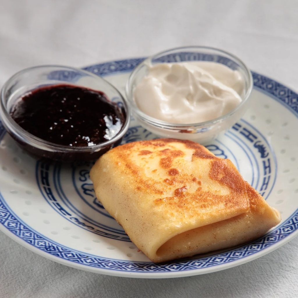 A photo of a blintz and two small condiment cups of sour cream and preserves beside it on a marble surface.