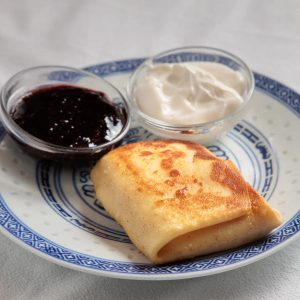 A photo of a blintz and two small condiment cups of sour cream and preserves beside it on a marble surface.