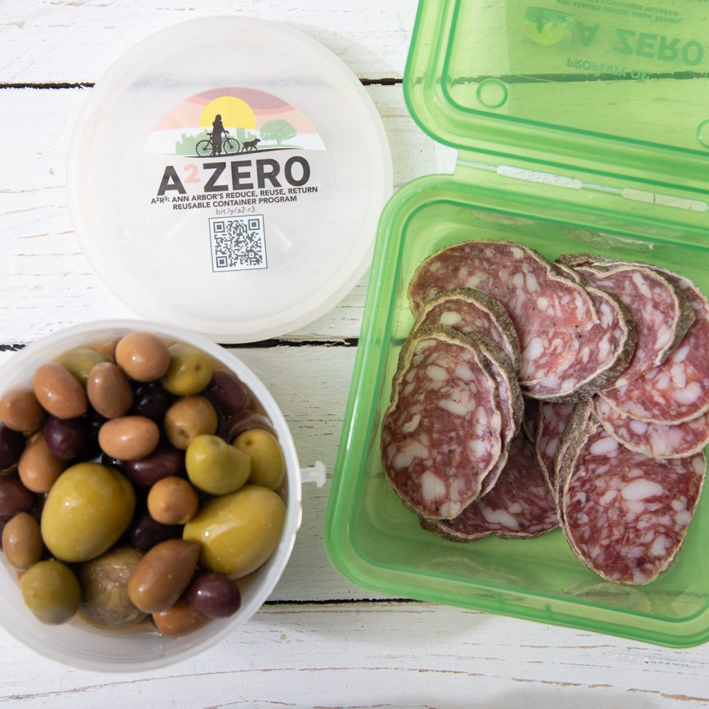 A top-down photo of two different A2Zero reusable containers, one filled with sliced salami and the other filled with an olive mix.