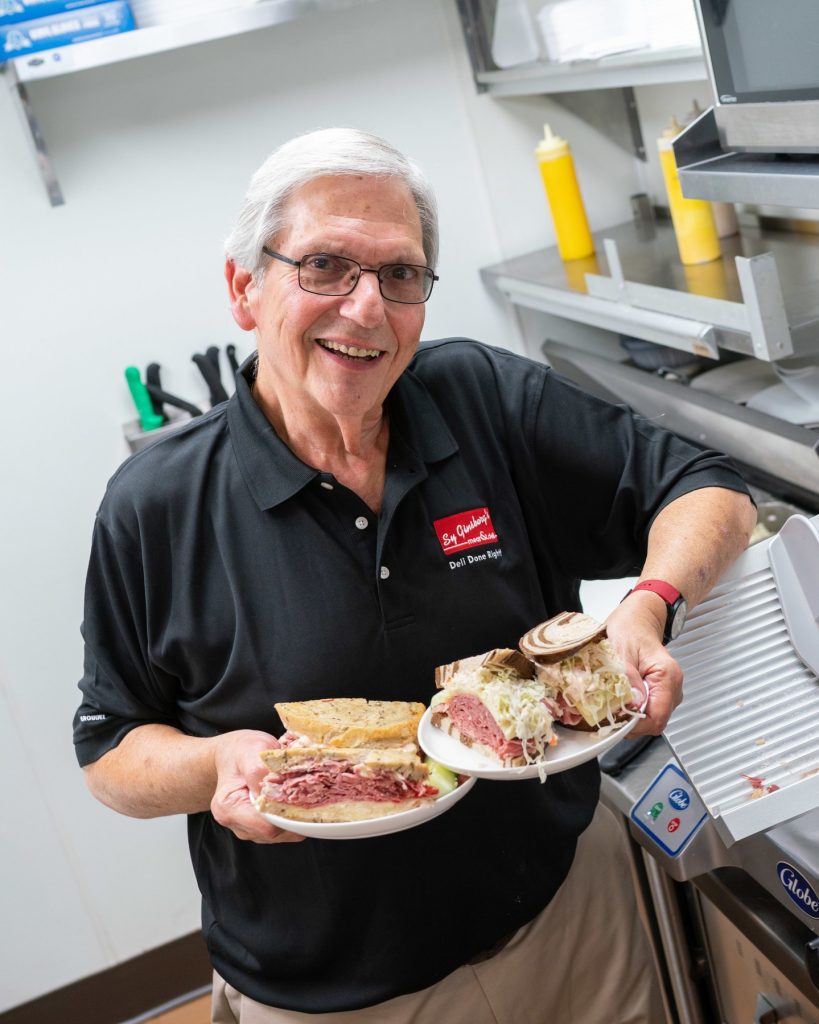 A photo of Sy holding two plates with corned beef sandwiches on them, smiling at the camera.