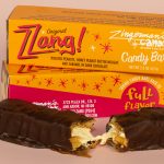 Zzang!® Bars Get Even Better Still! A photo of two Zzang bars in their packaging, with one unwrapped bar resting in front of them, broken in half to show the nuts and nougat inside.