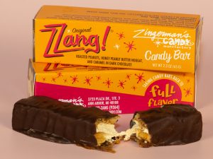 A photo of two Zzang bars in their packaging, with one unwrapped bar resting in front of them, broken in half to show the nuts and nougat inside.
