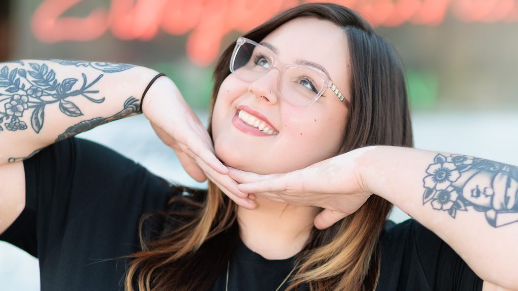 A portrait photo of Asha gazing upwards with a smile, and their fingers laced together beneath their chin. Asha has shoulder length brown hair that fades into blonde, a septum piercing, translucent square glasses, and tattoos down both their forearms.