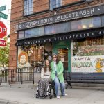 A photo of two women, one of whom is in a wheelchair, in front of the Deli building.