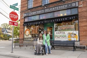 A photo of two women, one of whom is in a wheelchair, in front of the Deli building.