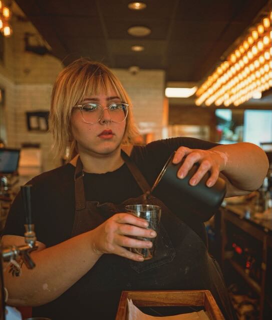 A photo of Asha working at a coffee shop, wearing a barista apron, pouring espresso into a cup.