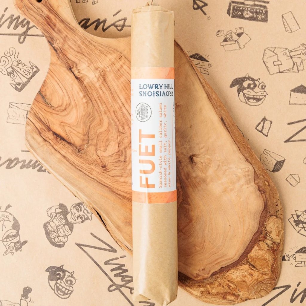 A top-down photo of a wrapped log of Fuet on a wooden serving board.