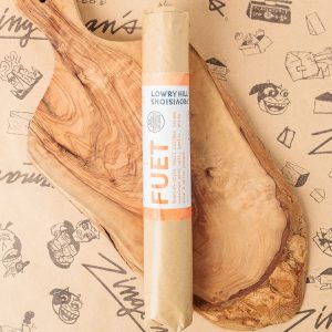 A top-down photo of a wrapped log of Fuet on a wooden serving board.
